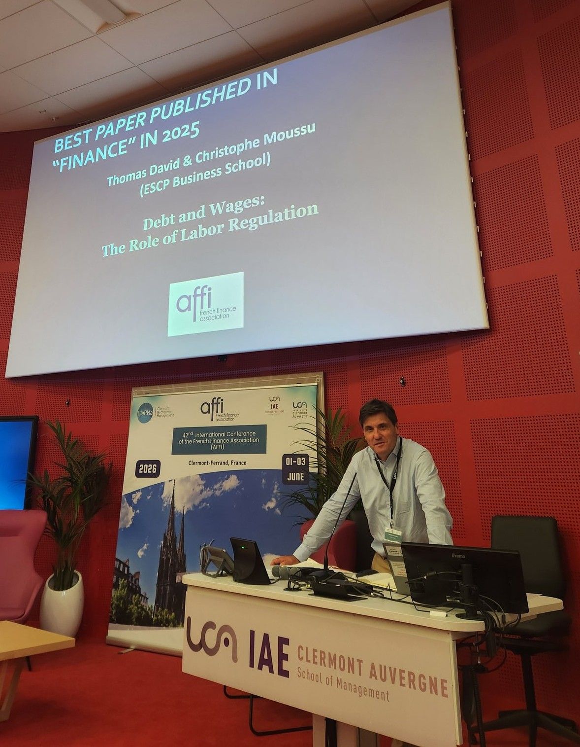 Professor Christophe Moussu stands at the podium during the French Finance Association (AFFI) conference as a screen displays the 2025 Best Paper Award for Debt and Wages: The Role of Labor Regulation by Thomas David and Christophe Moussu.

