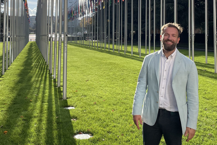 Marc Oberhauser standing among rows of national flags outside the UN building in Geneva on a sunny day.
