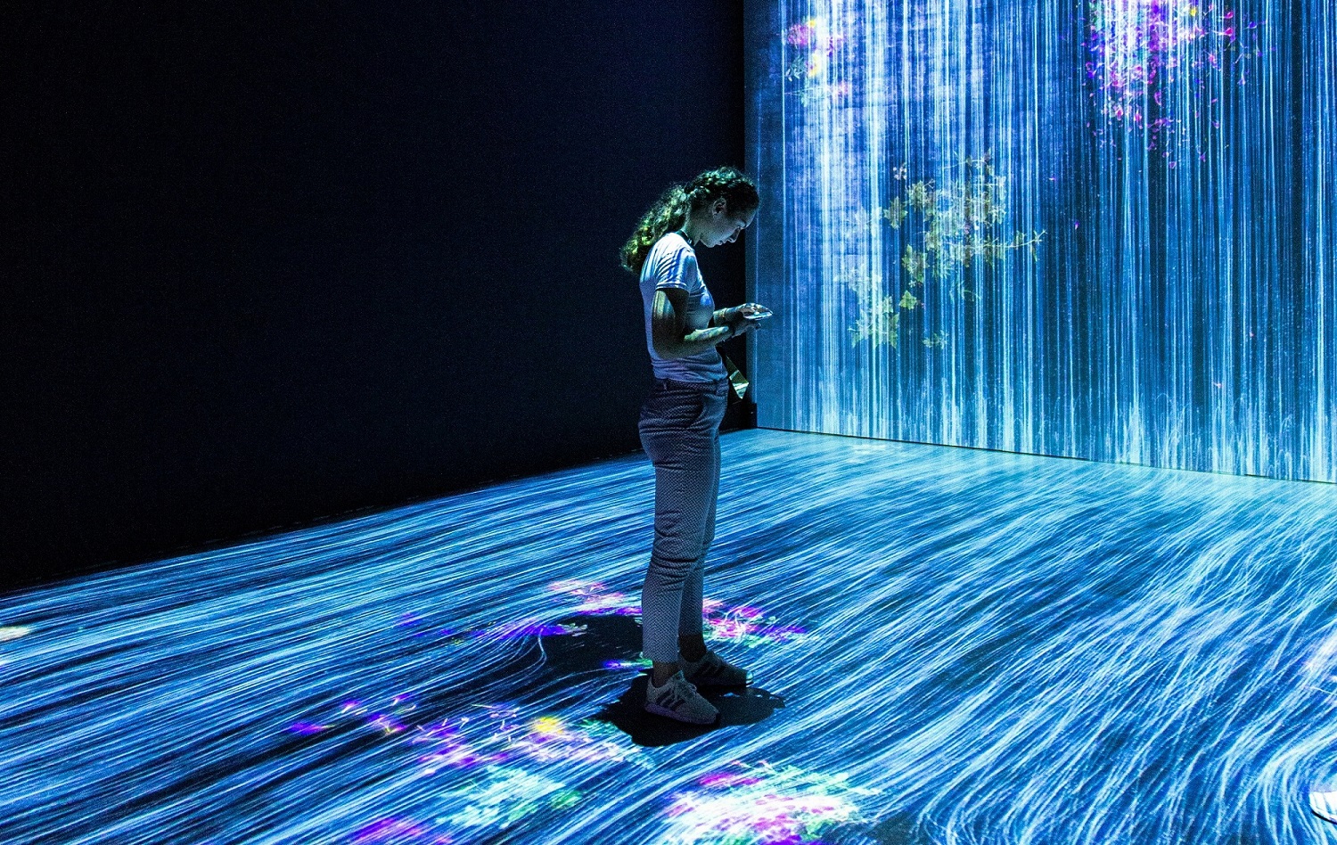 A woman stands in a dark room while light on screens flows from the wall and the floor.