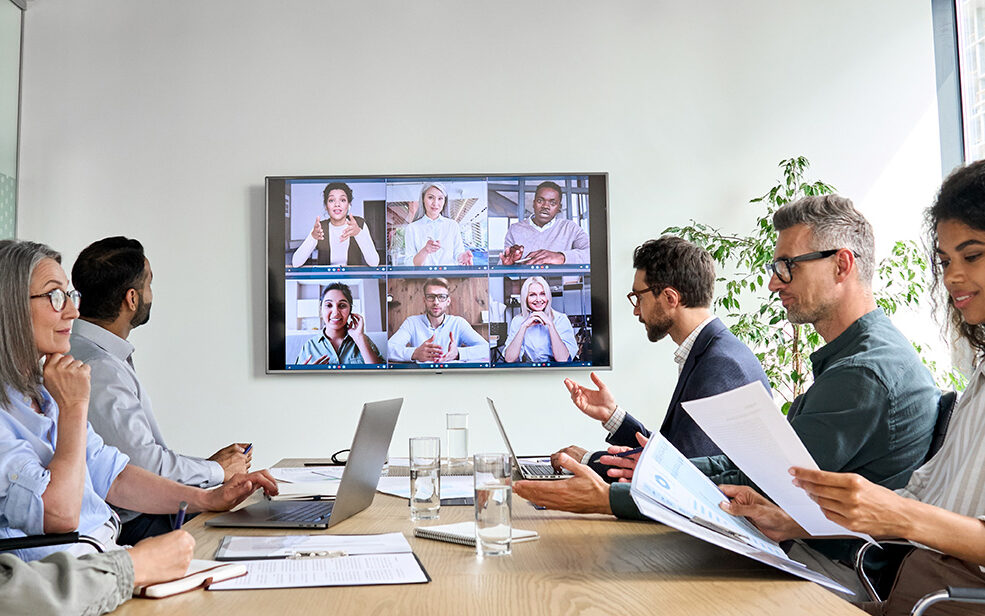 AdobeStock – insta_photos Diverse company employees having online business conference video call on tv screen monitor in board meeting room.