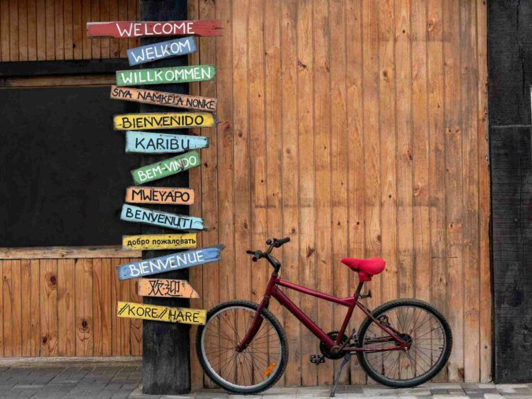 A red bike is propped up next to a tall sign that reads "welcome" in many languages.