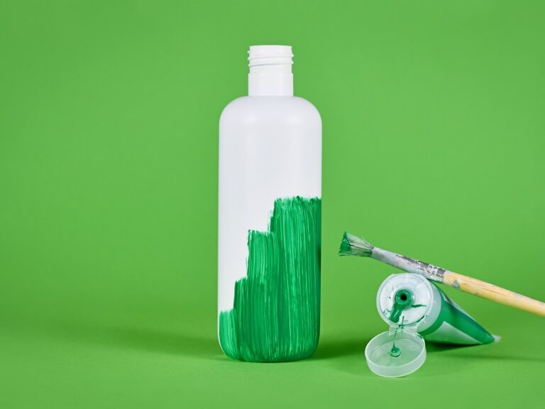 Photo of a white bottle on a green background, half painted by green paint with the paintbrush and open paint next to it.