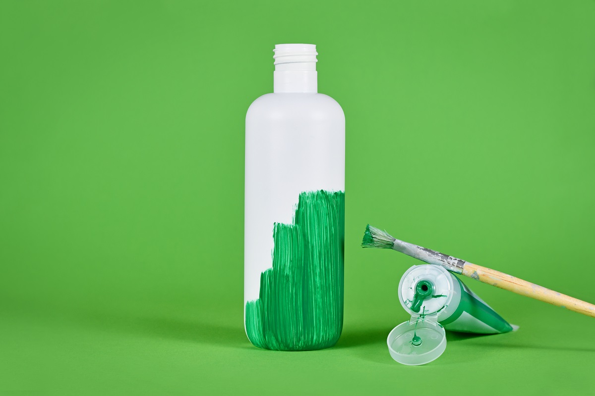Photo of a white bottle on a green background, half painted by green paint with the paintbrush and open paint next to it.