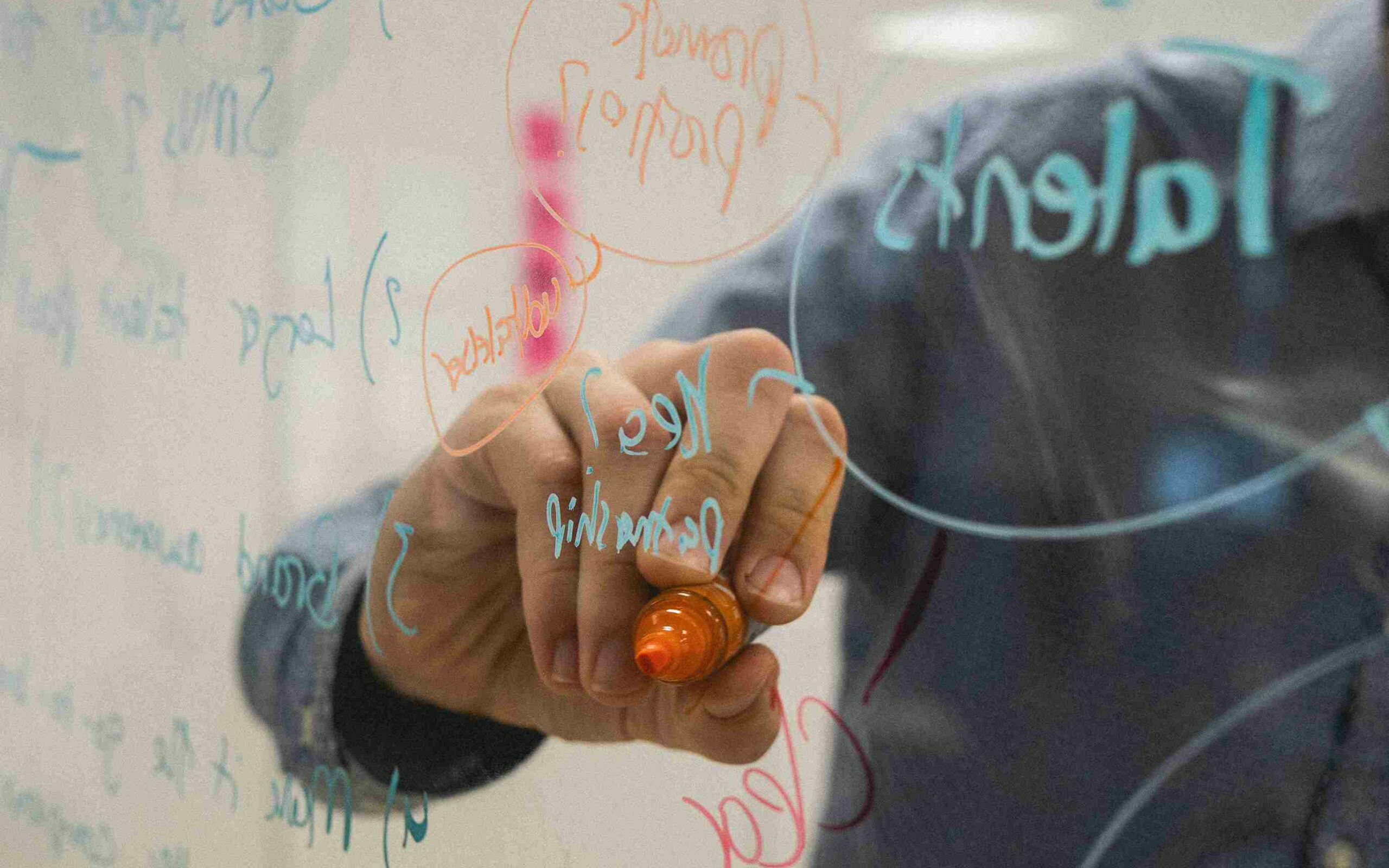 ©Kvalifik / Unsplash A man writes on a clear glass in what looks like an ideation session. The photo is taken from behind the glass.