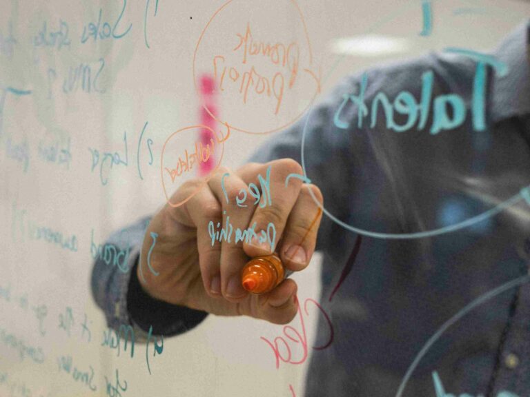 A man writes on a clear glass in what looks like an ideation session. The photo is taken from behind the glass.