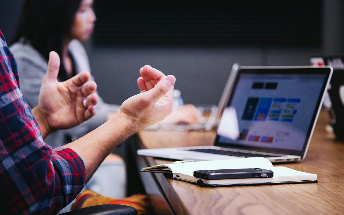 ©Headway / Unsplash Picture of a man sitting at a table with his laptop open in front of him. His arms are up as if in conversation and you can see a woman colleague in the background.