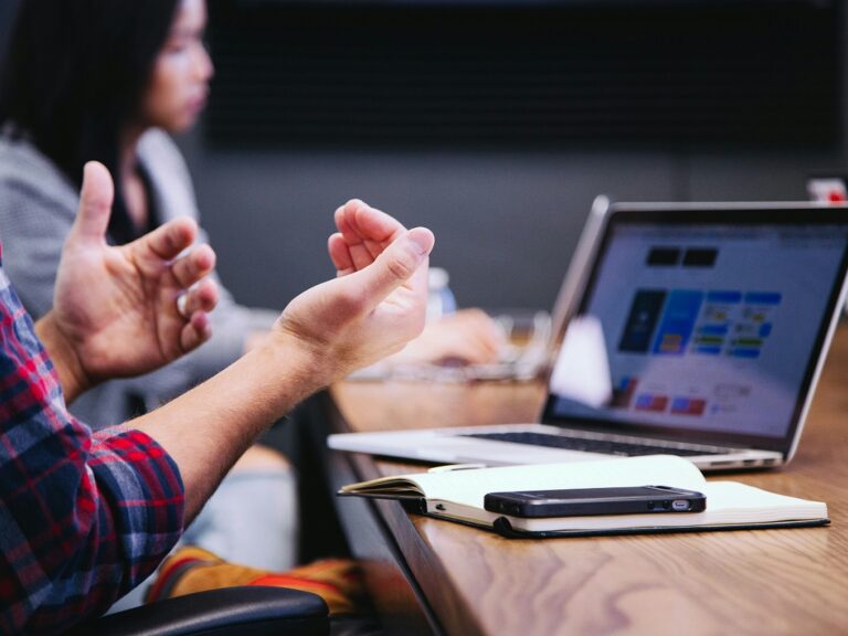 Picture of a man sitting at a table with his laptop open in front of him. His arms are up as if in conversation and you can see a woman colleague in the background.