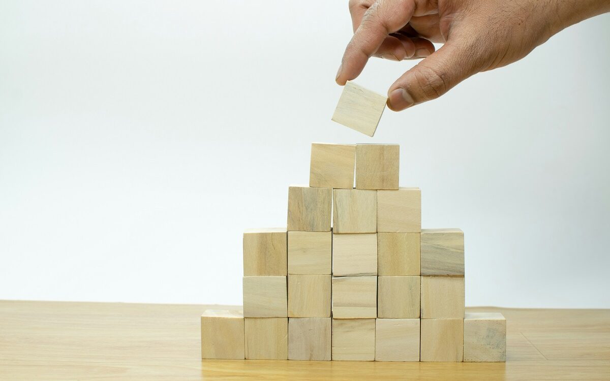 ©Imagine Buddy / Unsplash There is a pyramid constructed of 6 tiers of square wooden blocks. A hand is reaching in and taking off the top-most block of the pyramid.