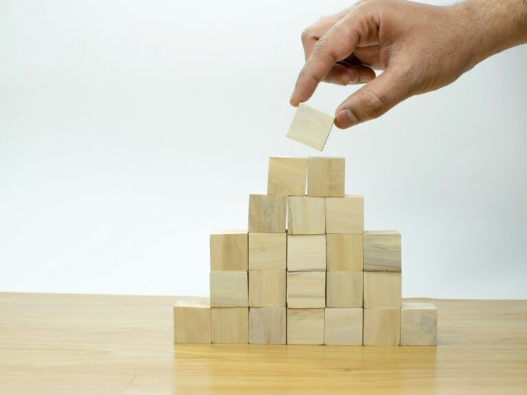 There is a pyramid constructed of 6 tiers of square wooden blocks. A hand is reaching in and taking off the top-most block of the pyramid.
