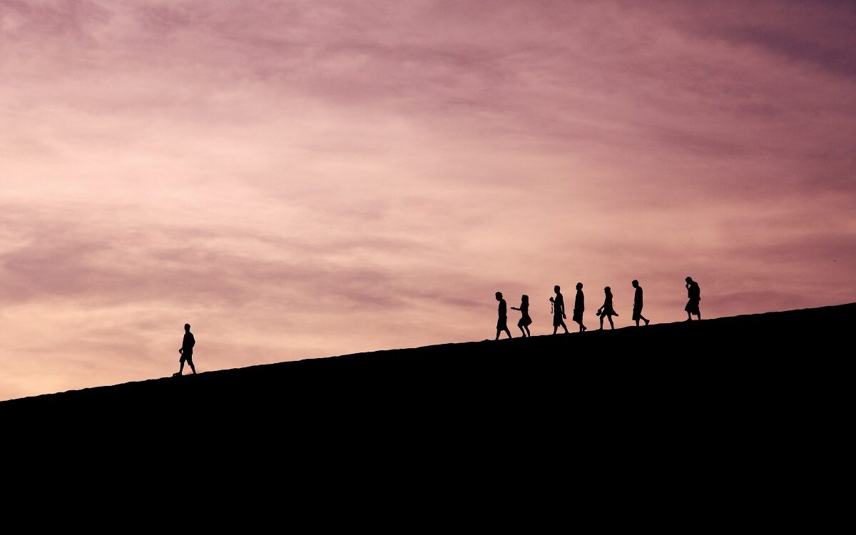 Silhouettes of people walking along the top of a hill. One person is out in front, leading the way.