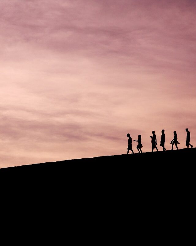 Silhouettes of people walking along the top of a hill. One person is out in front, leading the way.