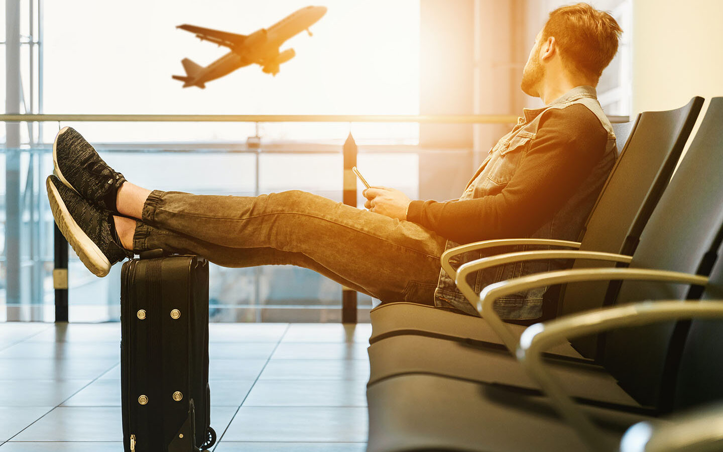 ©Jeshoot.com/Unsplash Man sitting on gang chair with feet on luggage looking at airplane,