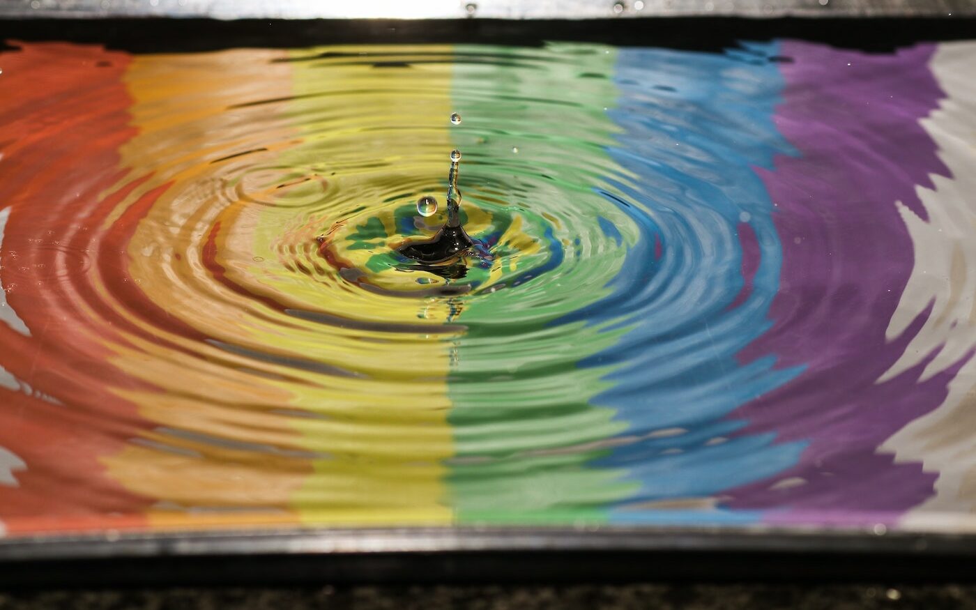 ©Jordan Mcdonald/Unsplash Reflection of a rainbow flag in water with a drop of water causing ripples.