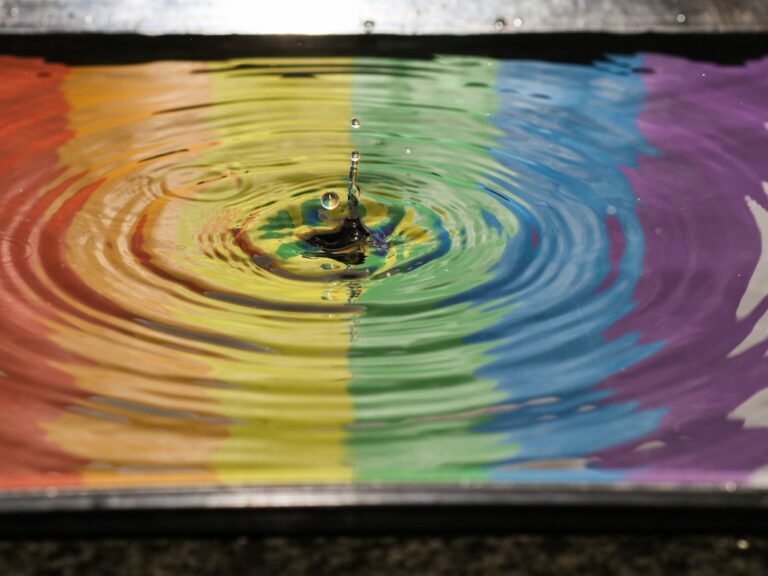 Reflection of a rainbow flag in water with a drop of water causing ripples.
