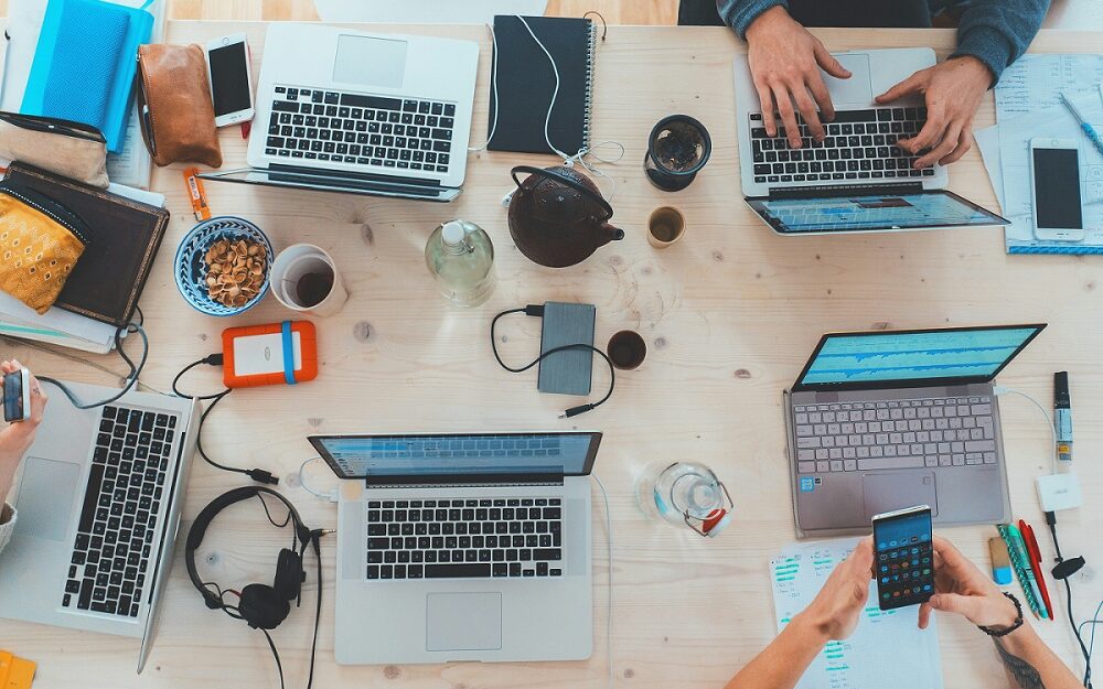 An overhead view of people working together on one big table showing their laptops, snacks, and hands.