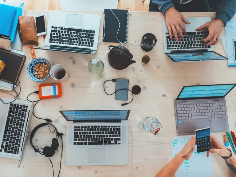 An overhead view of people working together on one big table showing their laptops, snacks, and hands.