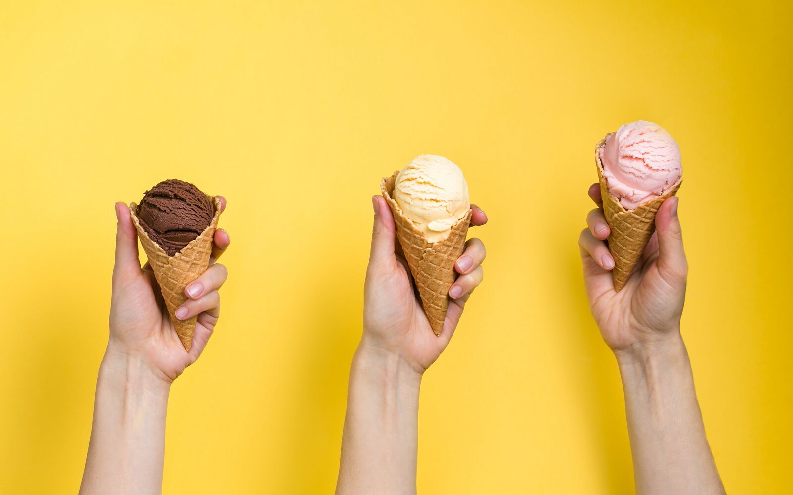 ©Oleksandra Naumenko / Shutterstock Three hands holding up three different ice cream cones with a scoop of different flavors of ice cream on a yellow background