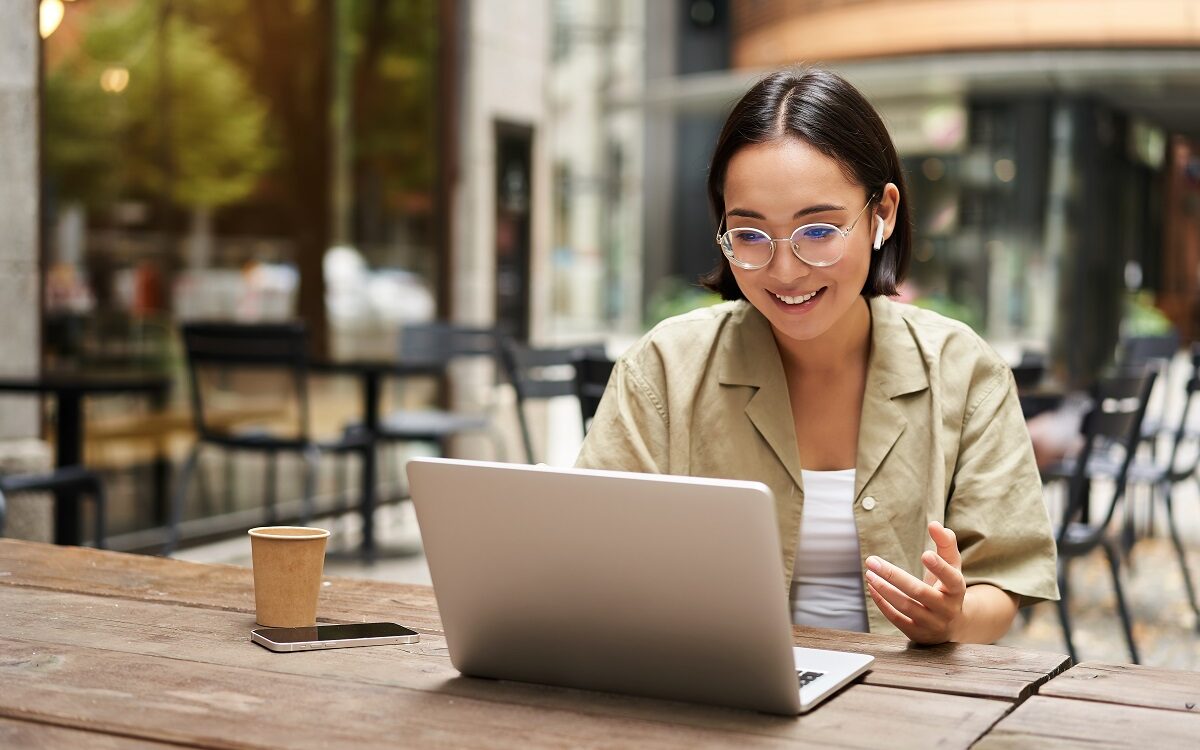 ©Mix and Match Studio / Shutterstock A woman sits in a cafe in front of an open computer, seemingly in the process of doing a video meeting
