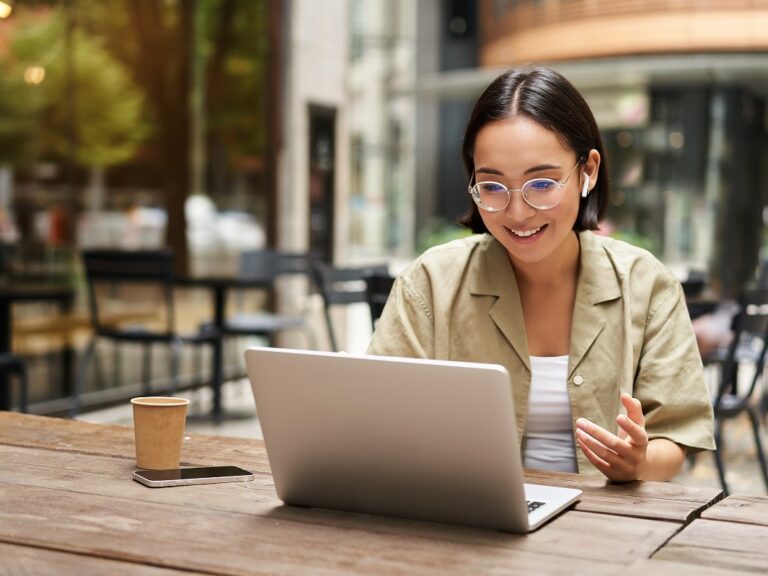 A woman sits in a cafe in front of an open computer, seemingly in the process of doing a video meeting