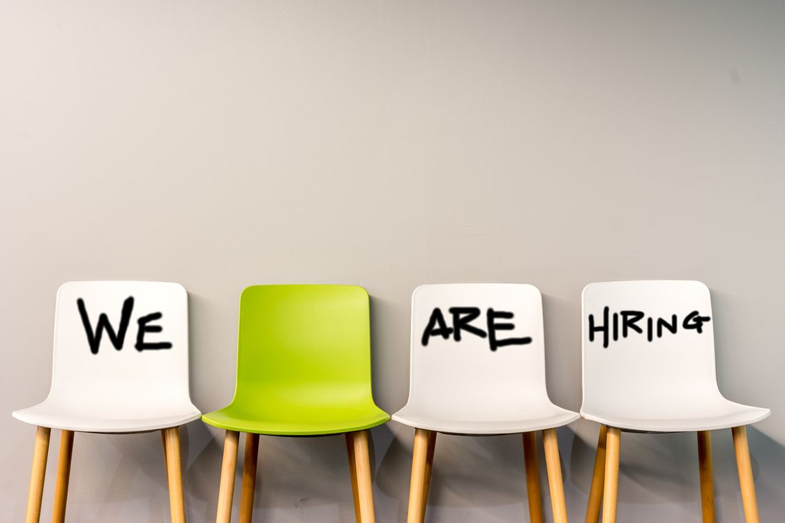 A row of 4 chairs with the writing "we are hiring" on the first, third, and fourth chairs which are white, but on the second chair, which is green it is left blank to signify that there is a seat availablein the company.