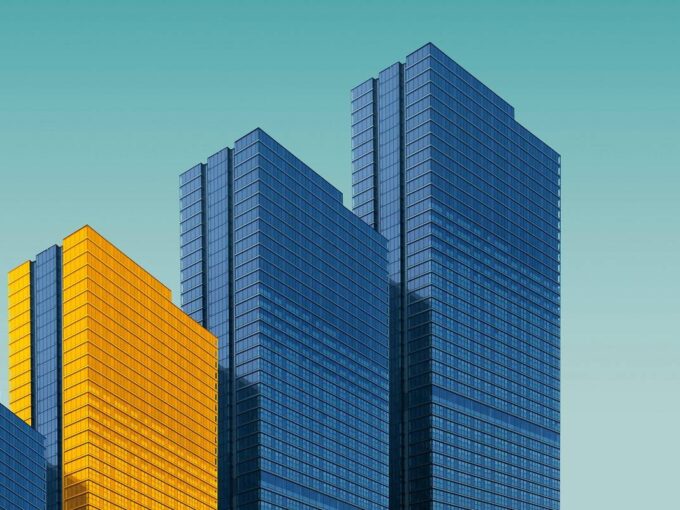 Four buildings in a row on a light blue background, three buildings are a normal grey color and one building is in yellow.