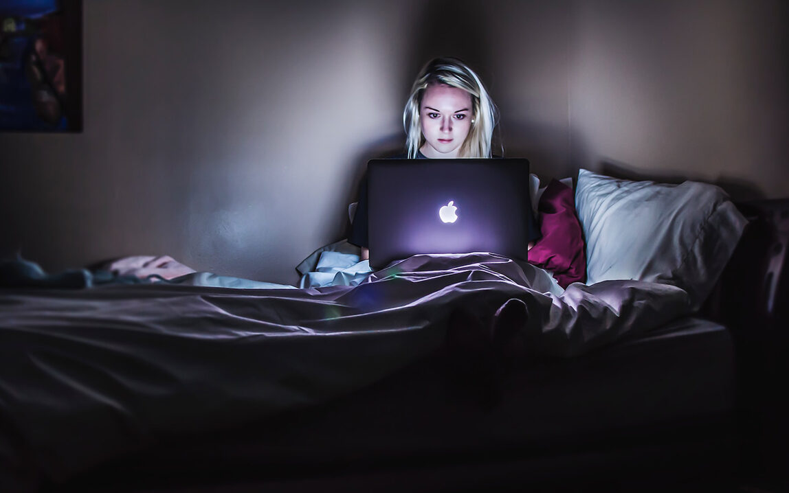 Victoria Heath – Unsplash Woman teleworking on a bed in the dark.
