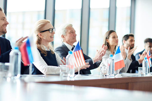A group of business people sitting at a table clapping with flags from different countries in front - by pressmaster / Adobe Stock